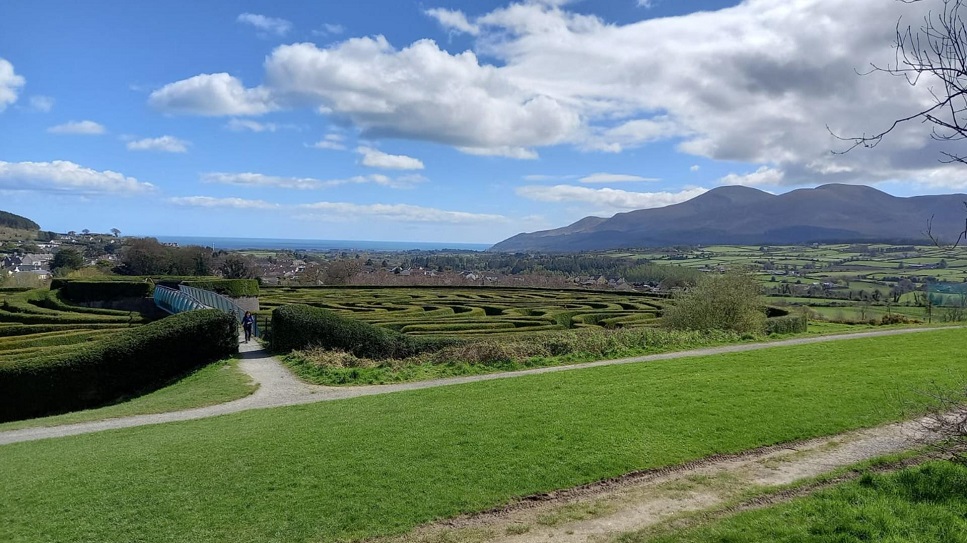 An image with two people walking out the exit of the Peace Maze in Castlewellan Forest Park, with the Mourne Mountains in the background. Photo taken on a bright day.